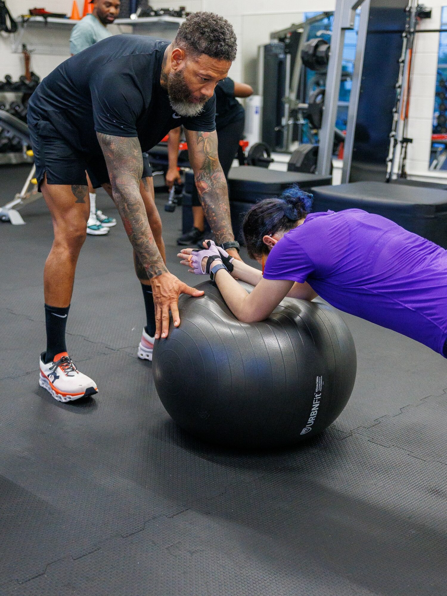 A man with tattoos and a beard is leaning over a large black exercise ball, assisting a woman in purple workout attire with an exercise.