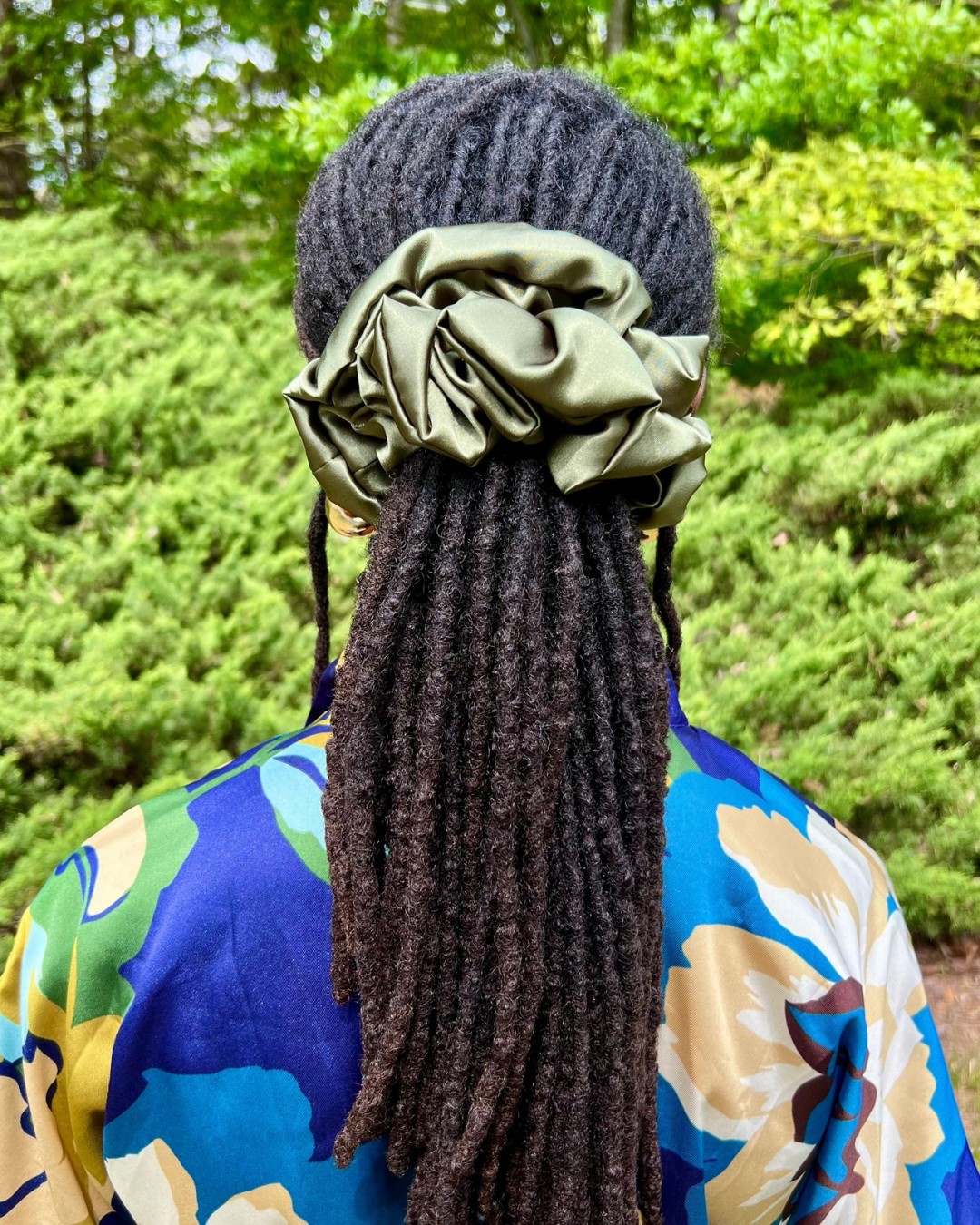 Person with long dreadlocks wearing a dark knit hat and large beige scrunchie, outdoors with green foliage in background.