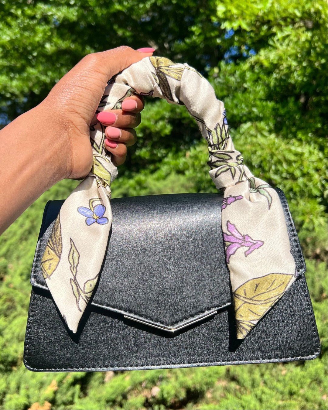 Hand holding a black handbag with a floral-patterned scarf handle against green foliage background.