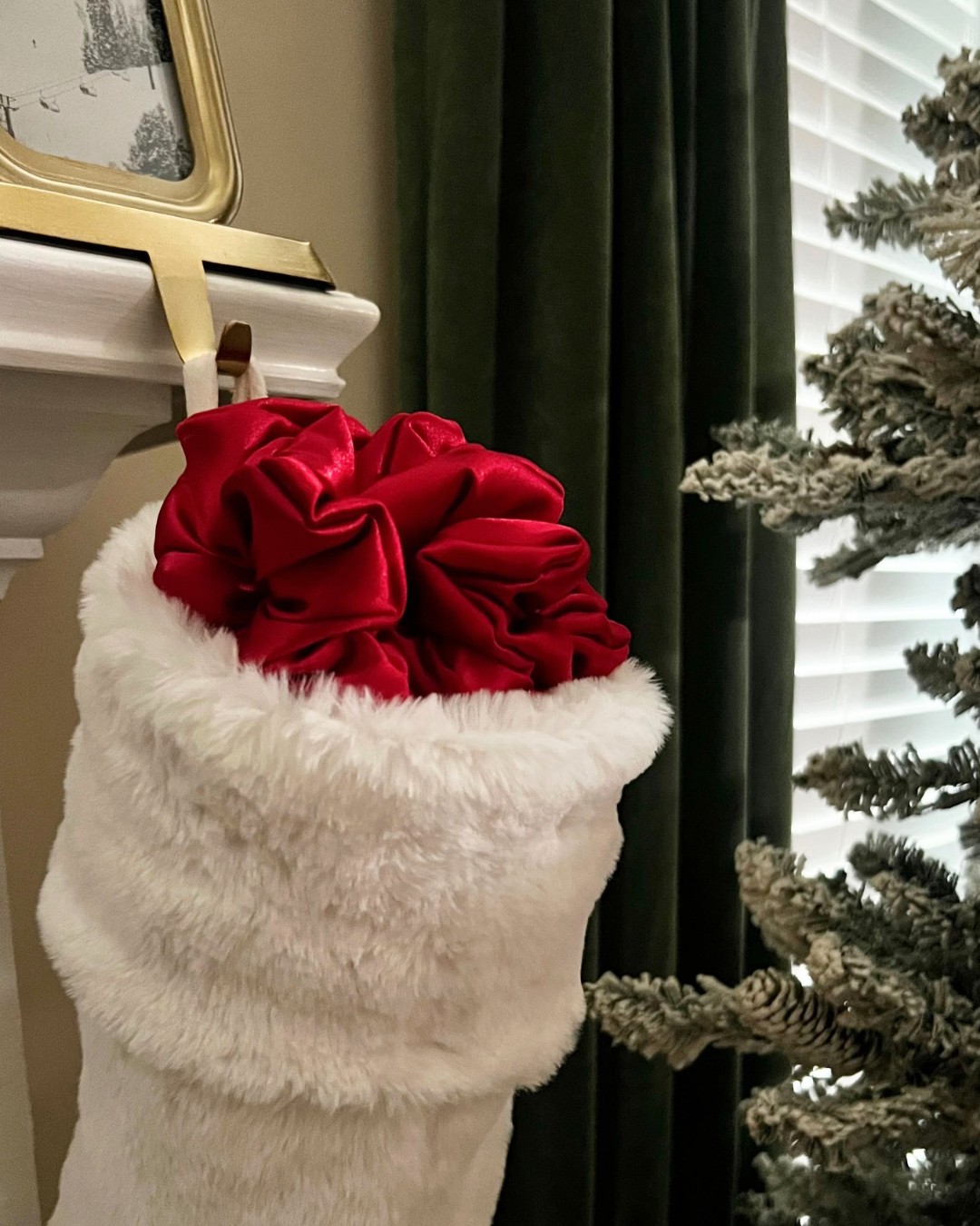 Red velvet bow on a white furry stocking hanging near a fireplace, with a Christmas tree and green curtains in the background.