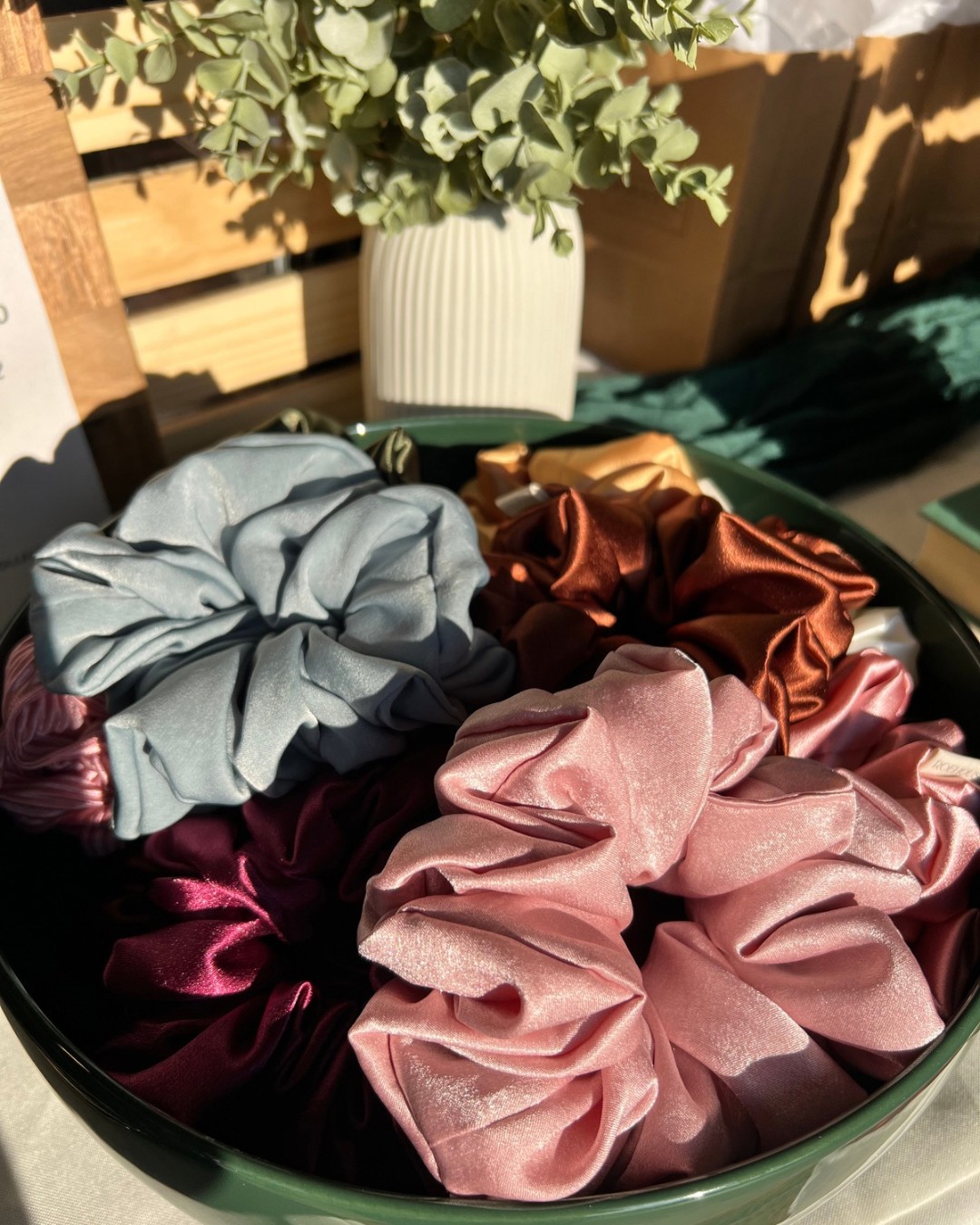 Colorful satin scrunchies in pink, burgundy, gray, and bronze inside a black container, with a potted plant in the background.