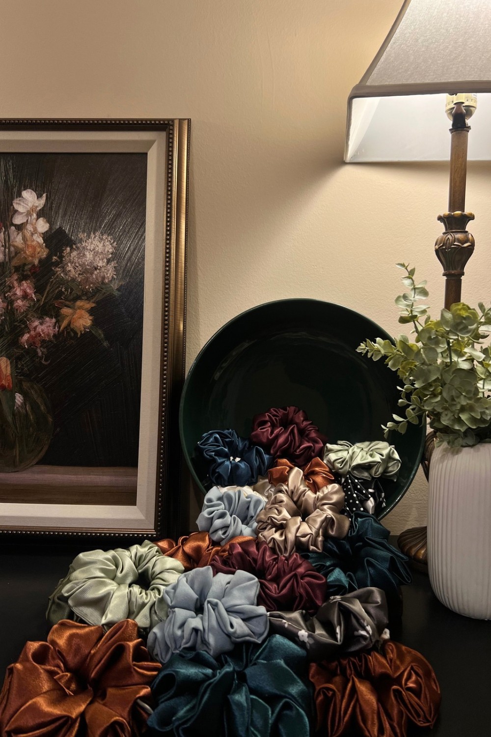 A table with a black tray holding colorful scrunchies, next to a white vase with green plants, a lamp, and a framed picture.