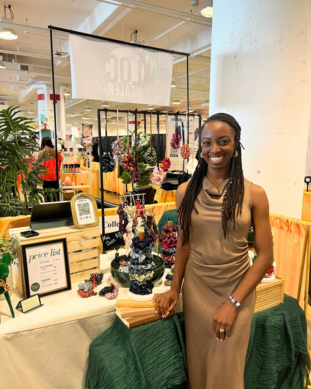Woman with long braids in a beige dress standing next to a display of jewelry and flowers at an indoor event.