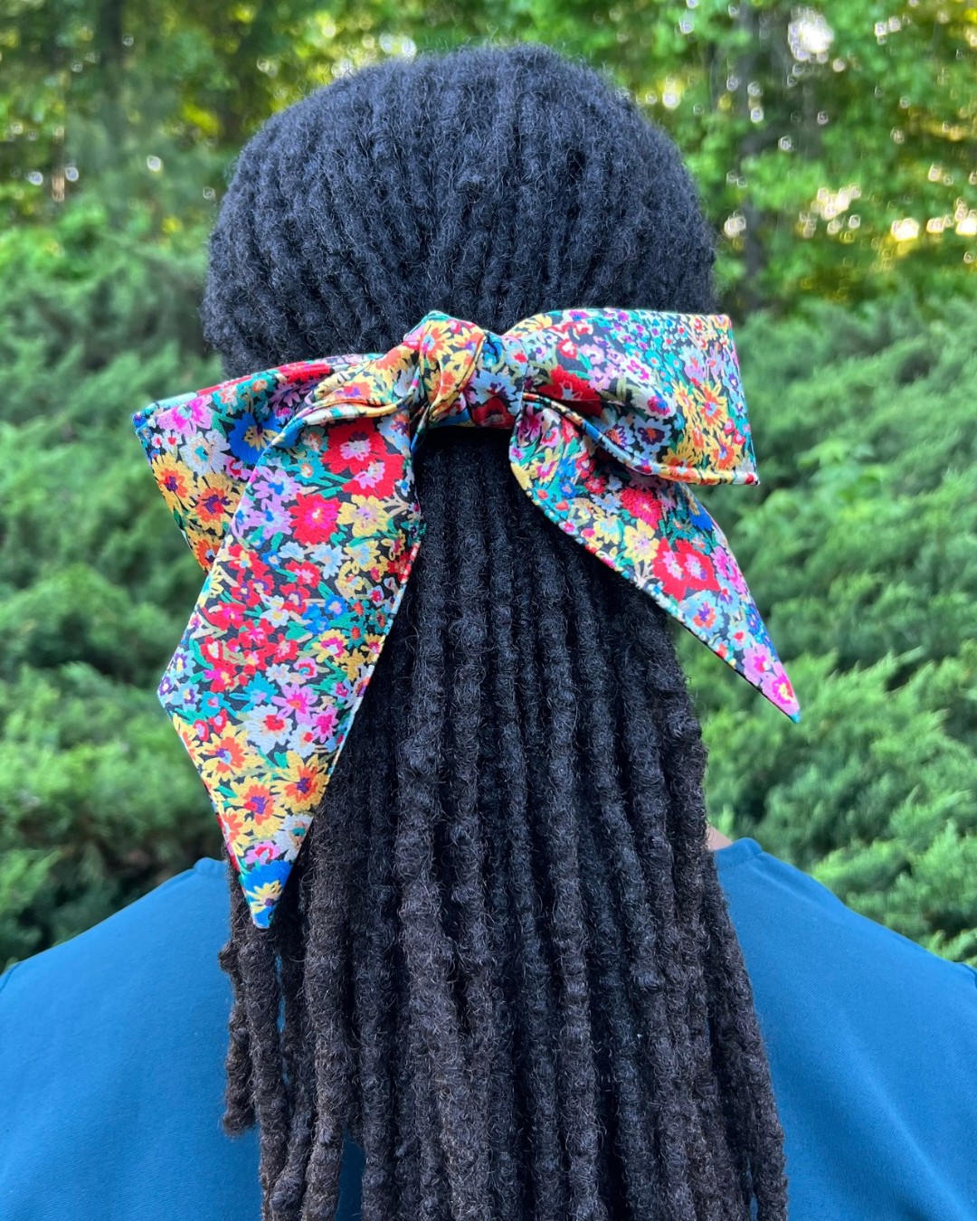 Person with long dreadlocks wearing a black knit hat and a colorful floral bow, outdoors with green trees in background.