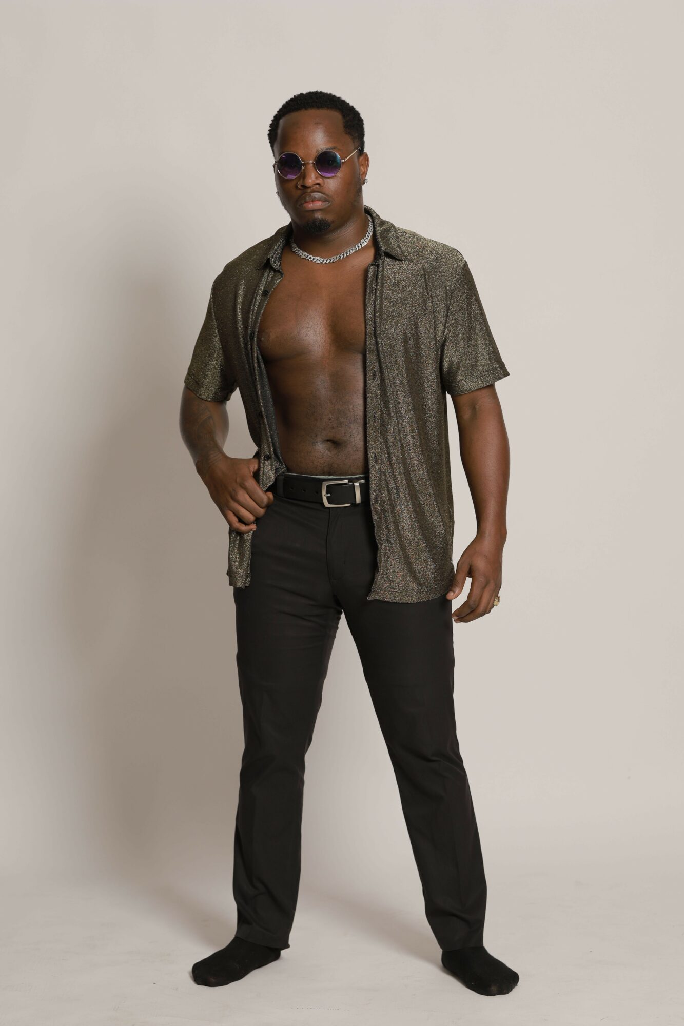 Man standing shirtless with sunglasses, wearing black pants and a gray jacket, against a plain background.