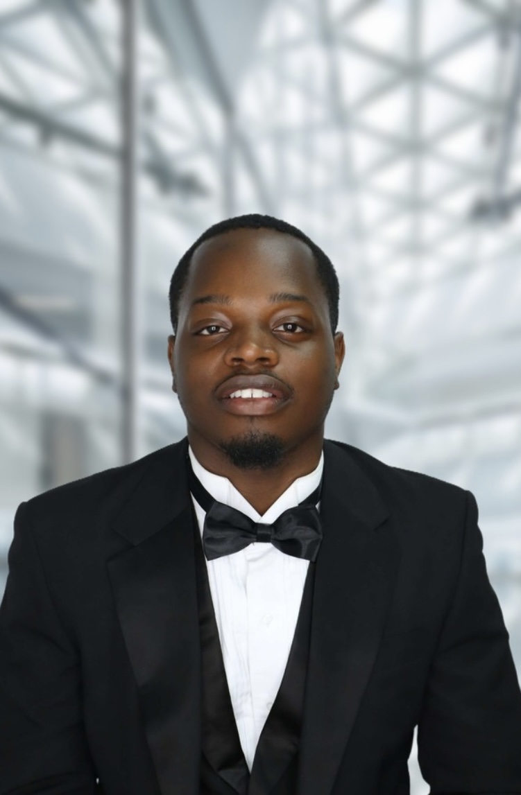 Man in tuxedo with bow tie standing indoors with a modern glass ceiling background.