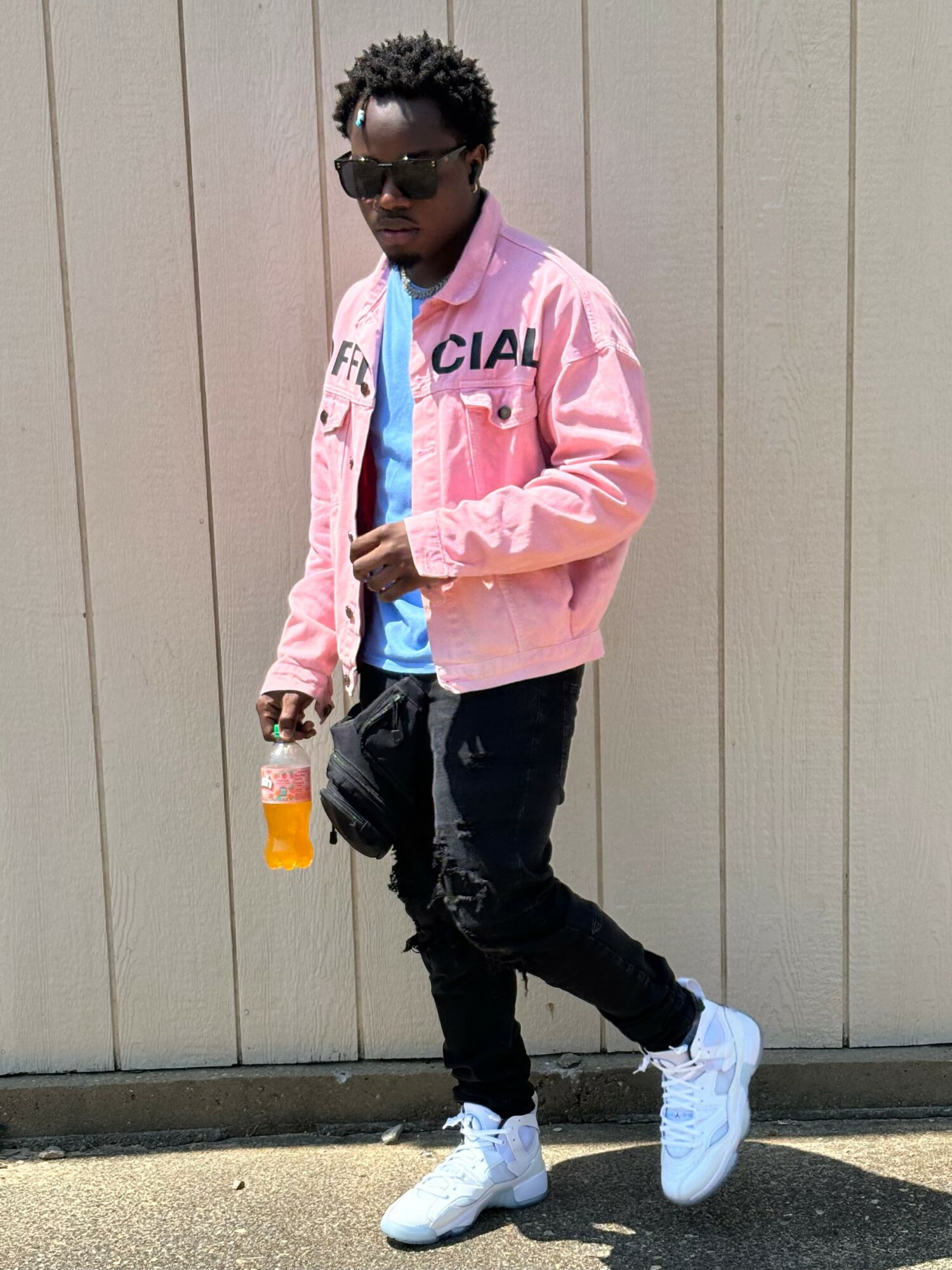 Young man with sunglasses, wearing a pink jacket, black pants, and white sneakers, holding a drink, standing outdoors against a beige wall.