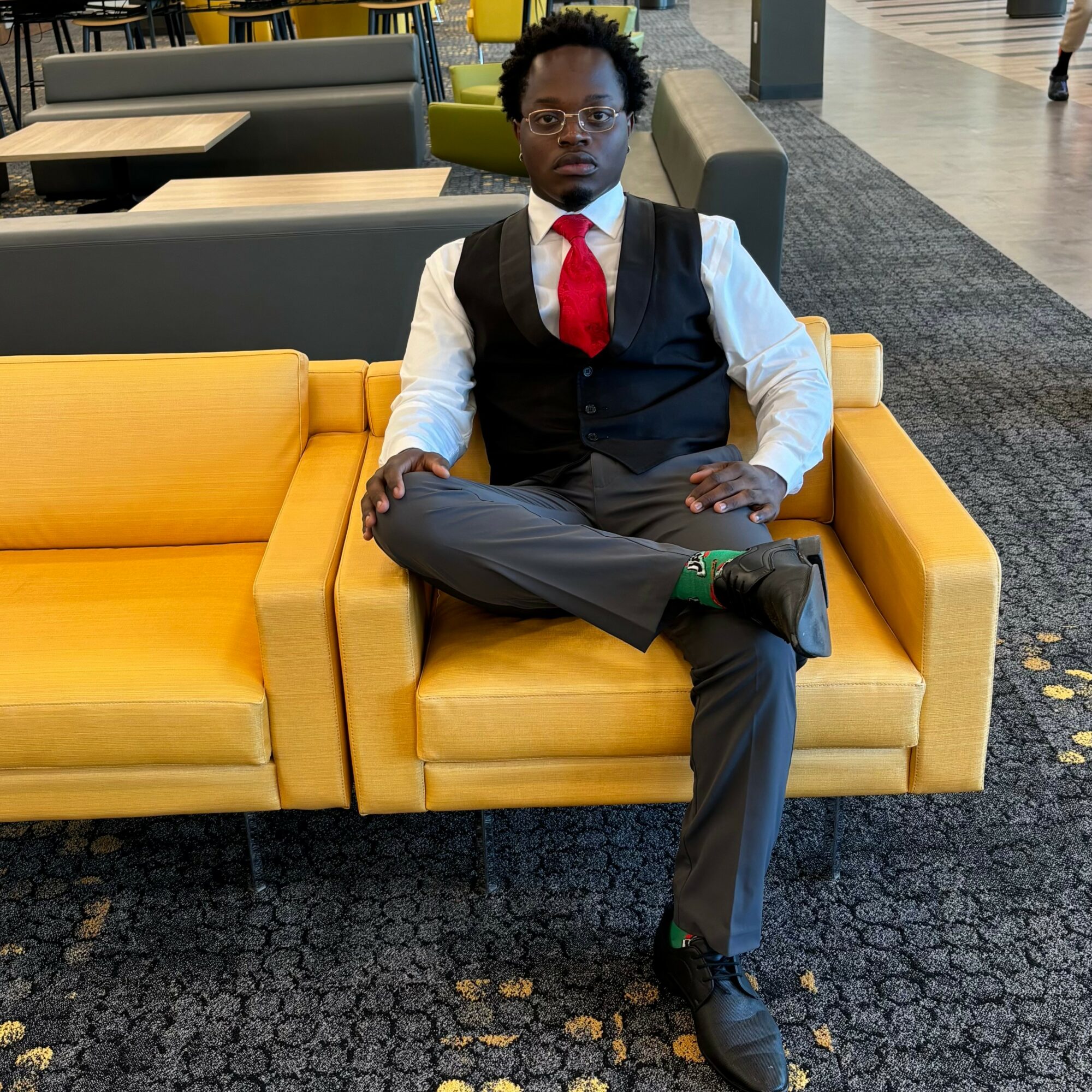 Man in formal attire sitting on a yellow sofa in a public space, with arms crossed and legs crossed.