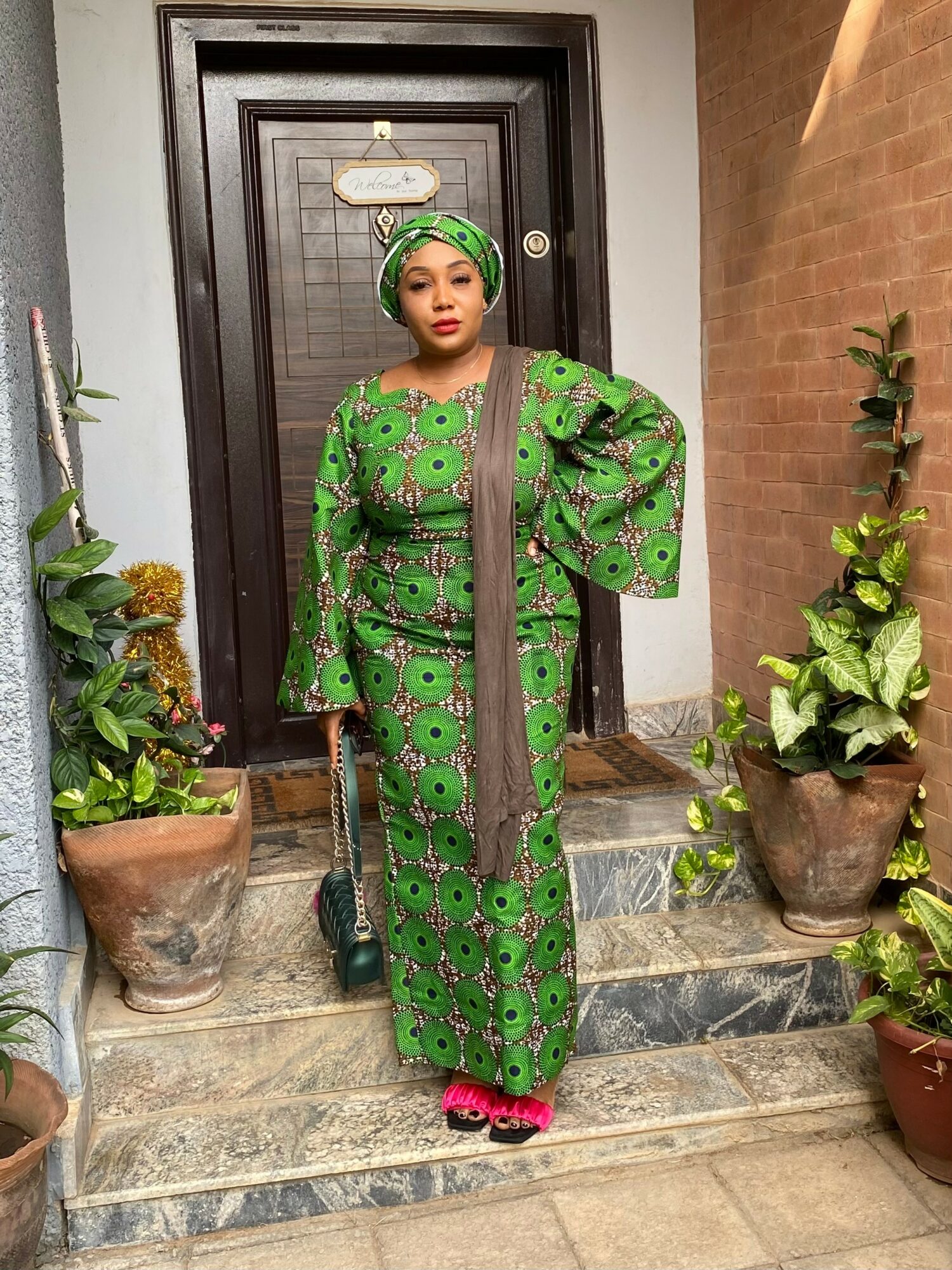 Woman in green traditional attire standing on steps with potted plants, door behind her, looking at camera.