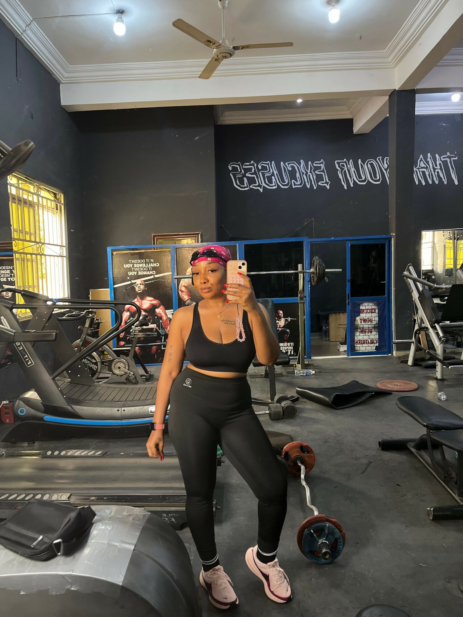 Woman in workout clothes standing in a gym with equipment and a chalkboard, looking at the camera.