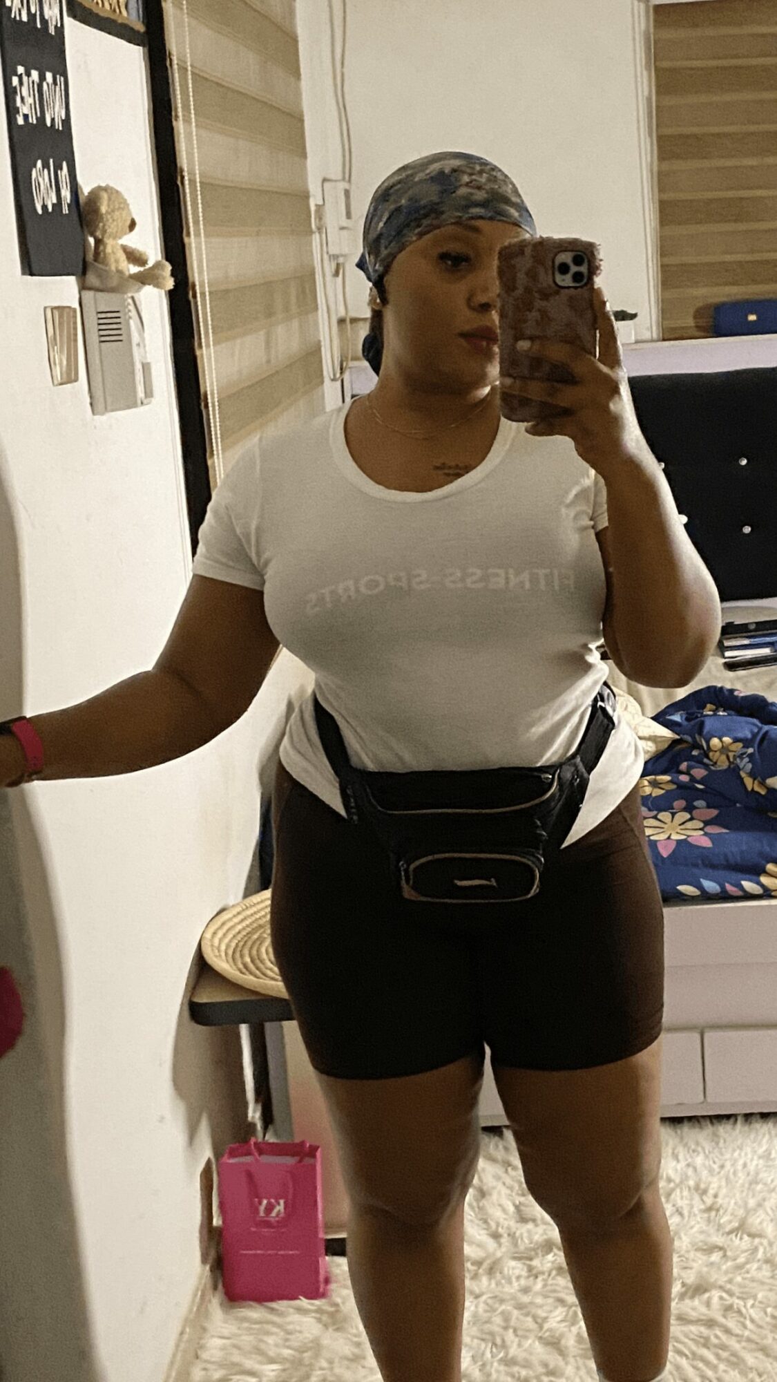 Person taking a mirror selfie in a room, wearing a white t-shirt, black shorts, and a headscarf, holding a phone.
