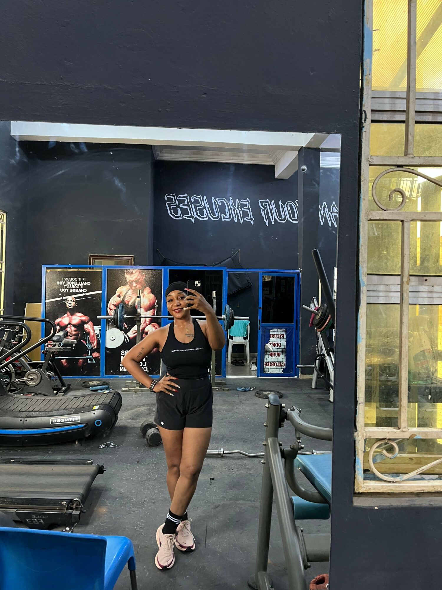 Two women stand in a gym, one holding a phone, with gym equipment and a blue shelf behind them.