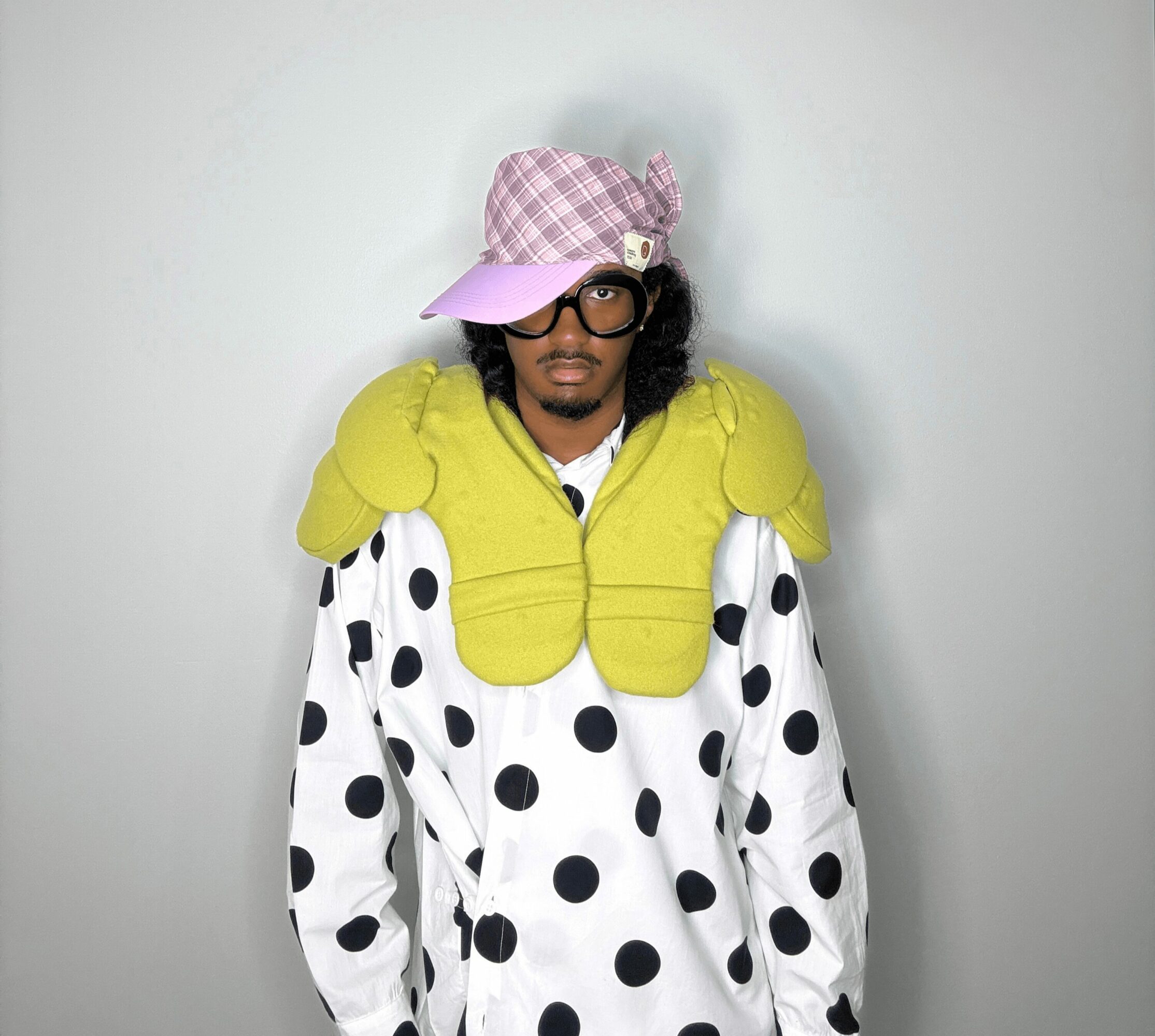 Person wearing a pink checkered hat, large glasses, a yellow padded shoulder piece, and a polka dot shirt, standing against a plain background.