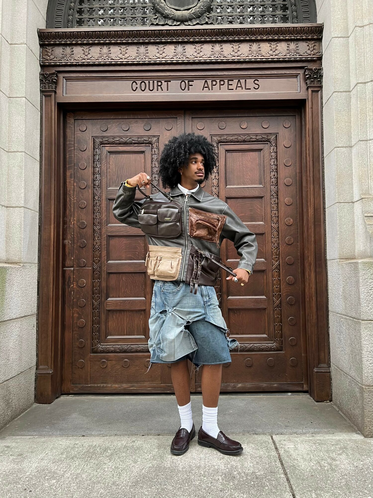 Person with curly hair standing in front of a wooden door labeled 'Court of Appeals', holding a rifle, wearing layered clothing and shorts.