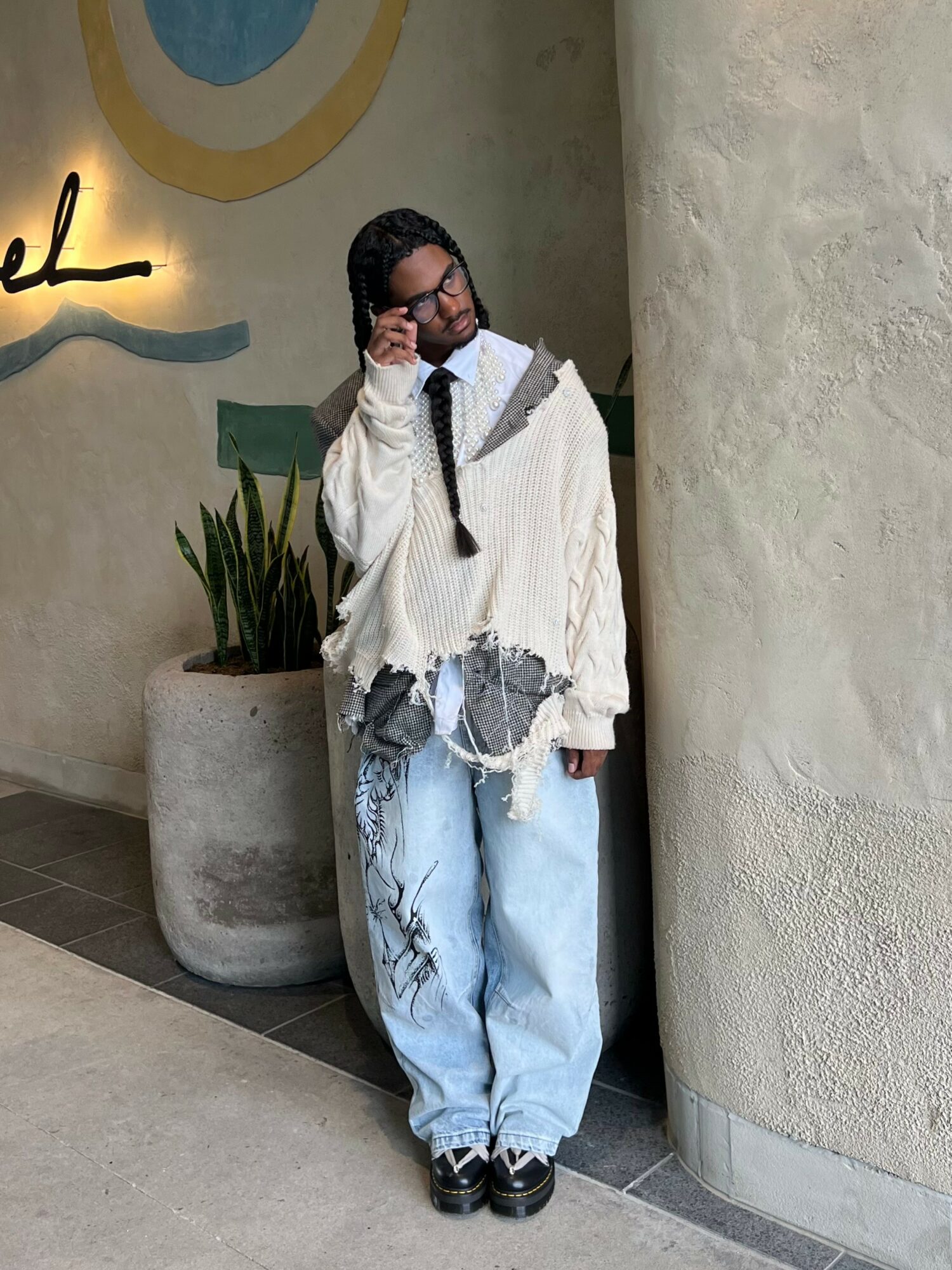 Person leaning against a wall, wearing glasses, a white hoodie, and baggy jeans, with a plant nearby.