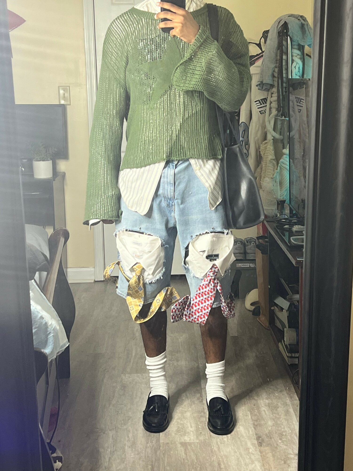 Person standing in a room wearing a green coat, white sweater, denim shorts, and black shoes, holding a bag.
