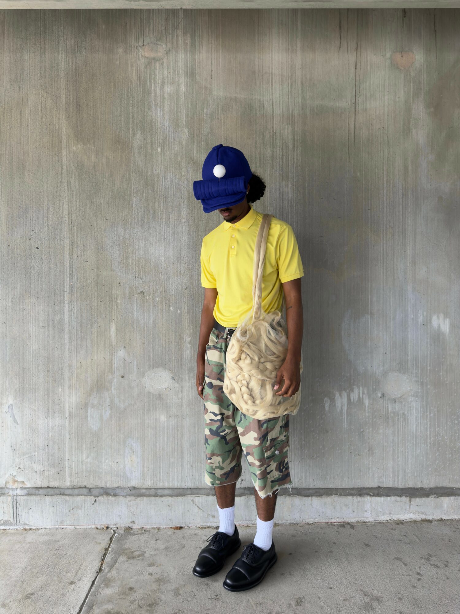 Person standing against a plain wall, wearing a blue hat, yellow shirt, camouflage pants, black shoes, and carrying a beige bag.