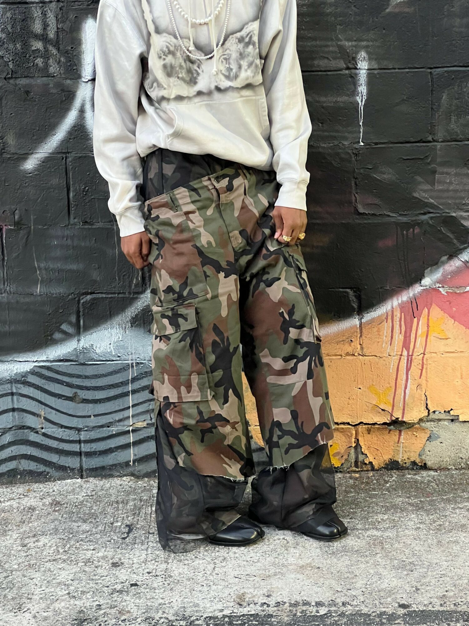 Person wearing camouflage pants and black shoes standing against a graffiti-covered wall.