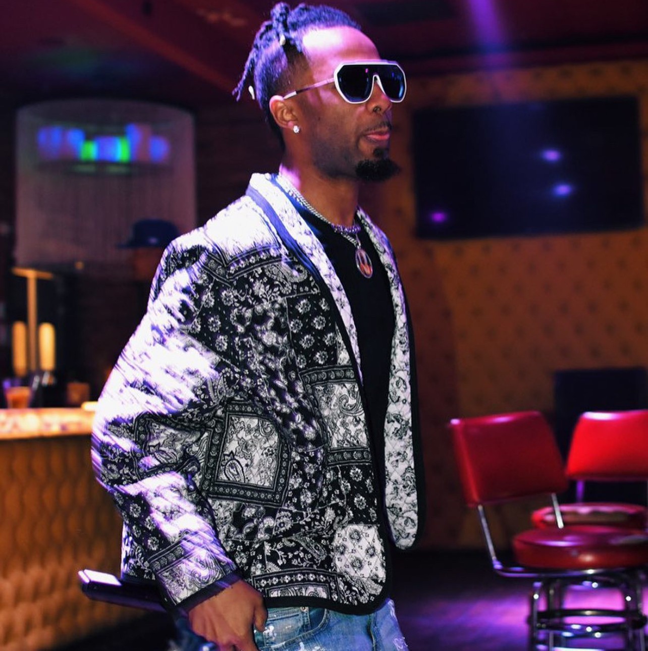 Man with sunglasses and patterned jacket in a dimly lit room with red chairs.