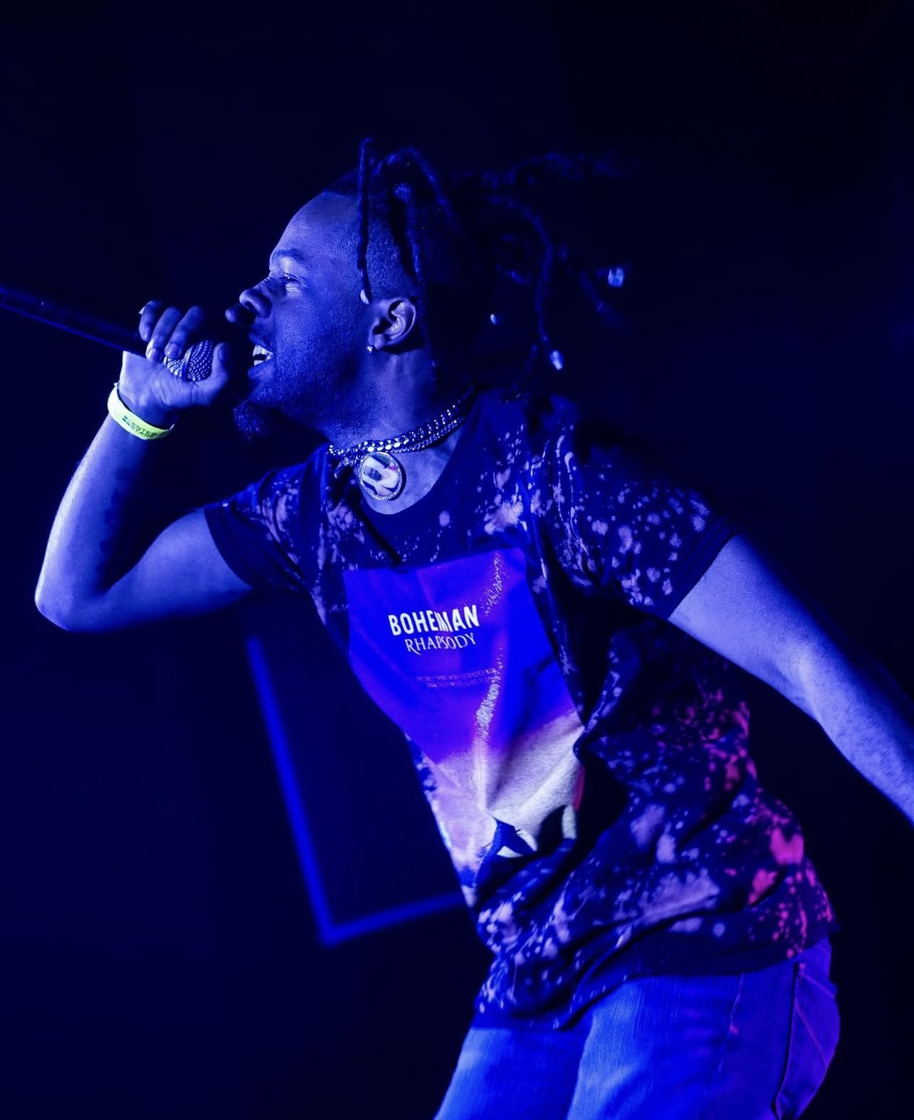 Person singing into a microphone on stage, wearing a galaxy-patterned shirt and jewelry, with dark background.