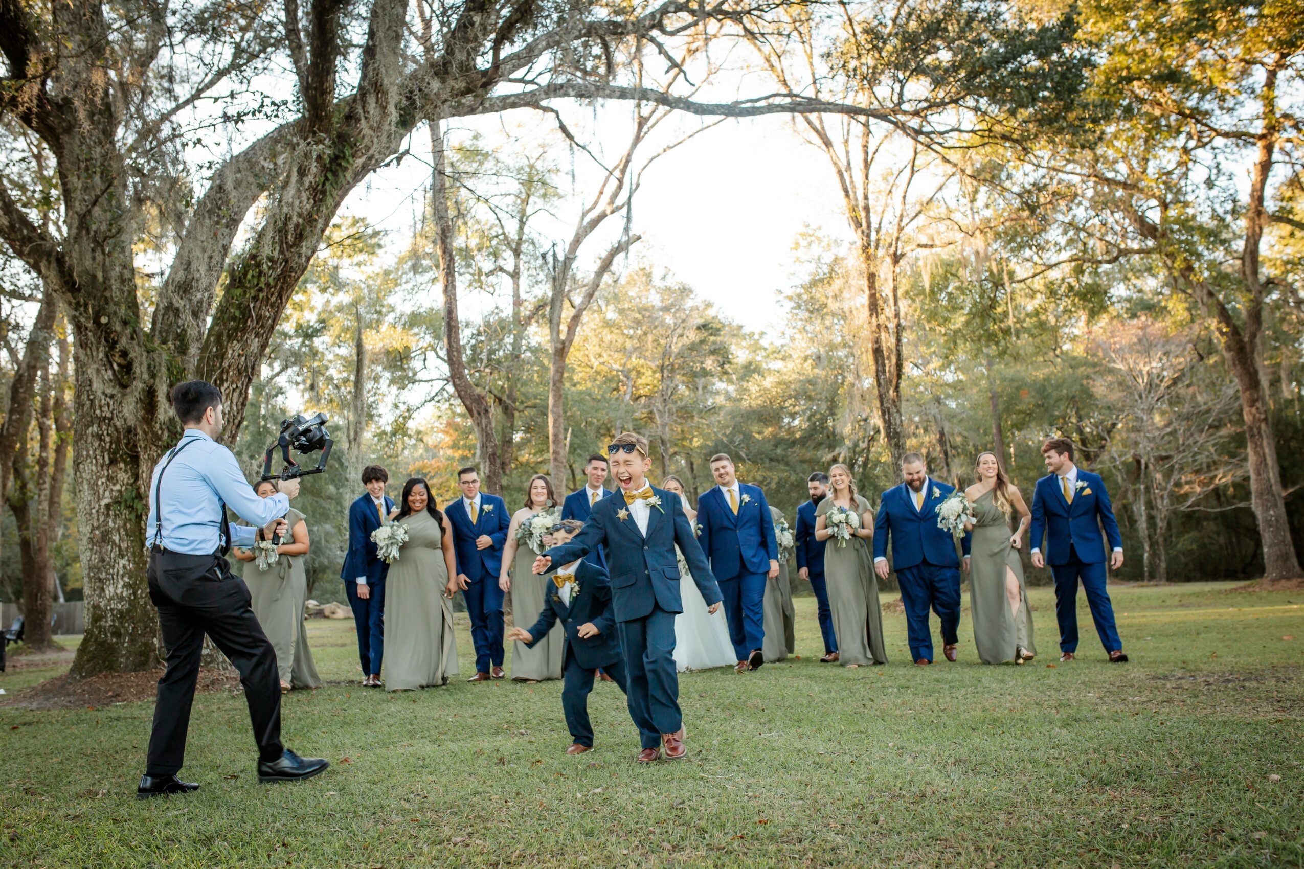 Group of people outdoors, some in formal attire, standing on grass with trees in background, sunlight filtering through leaves.