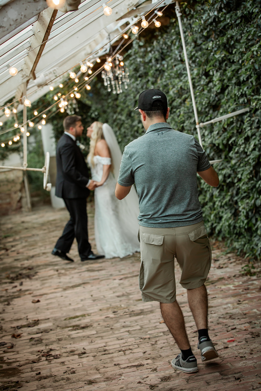 Photographer capturing a wedding couple standing outdoors under string lights, with a green hedge background, on a brick path.
