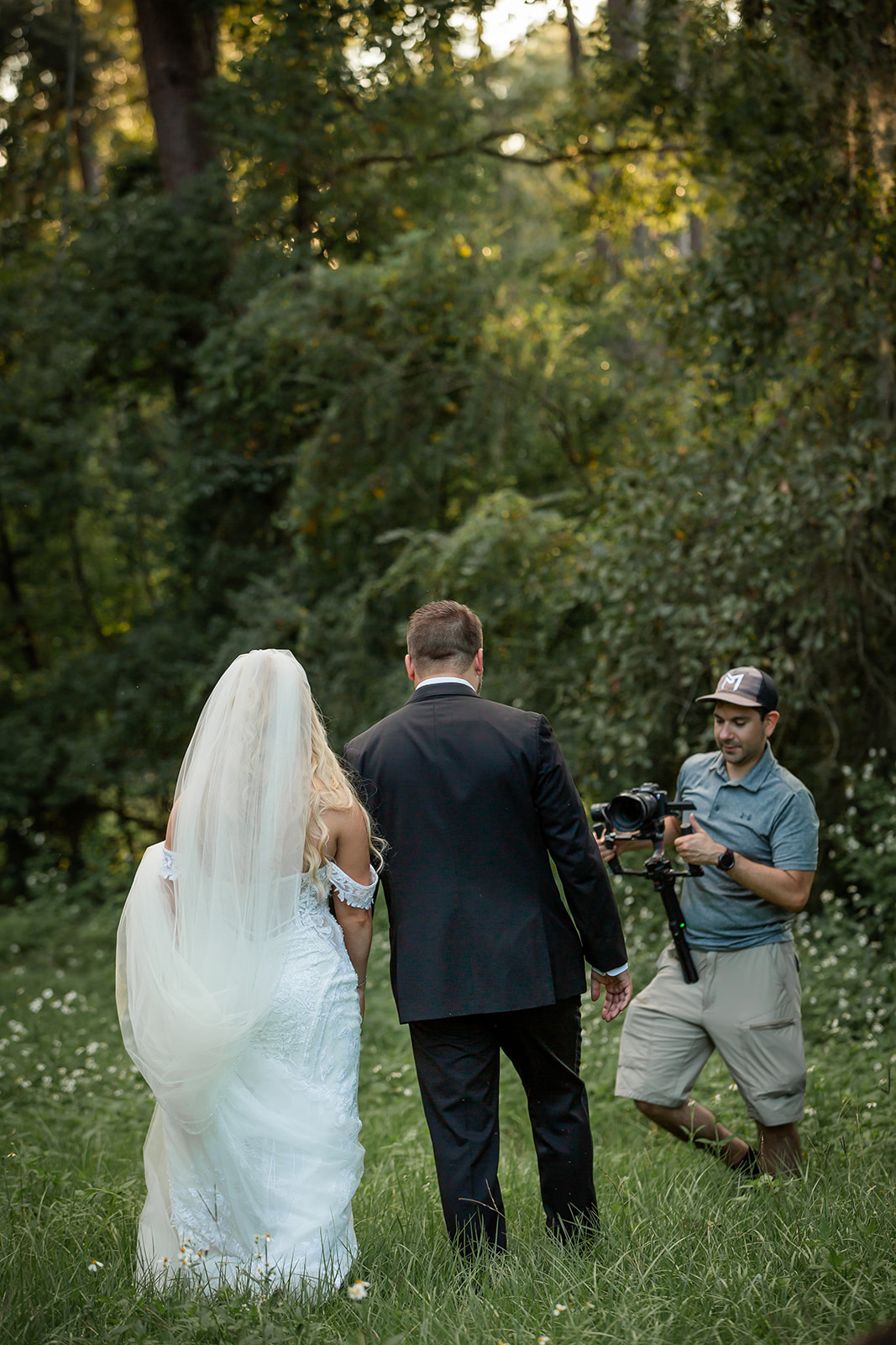 A bride and groom stand outdoors on grass, with a photographer capturing the moment, surrounded by trees and sunlight.