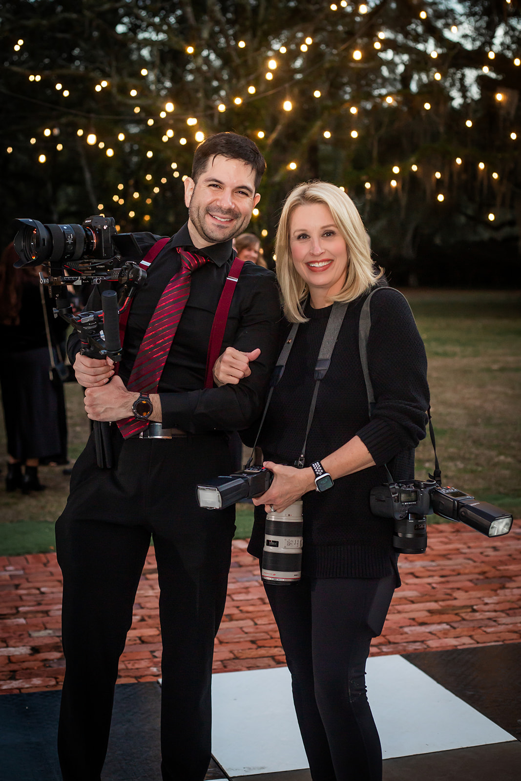 Two photographers, a man and a woman, smiling outdoors at night with string lights in the background, holding cameras.