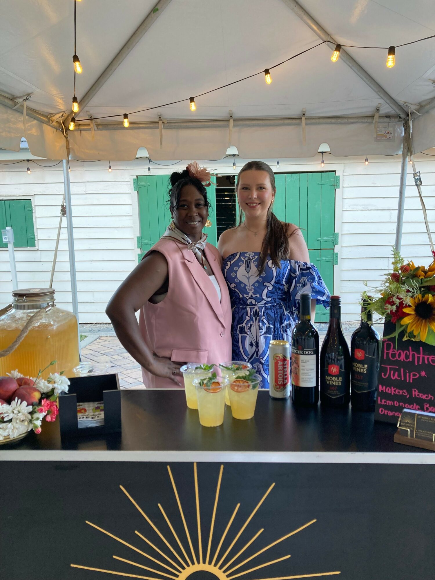 Two women standing behind a bar with drinks, flowers, and bottles, under a tent with string lights, green shutters, and a sign.