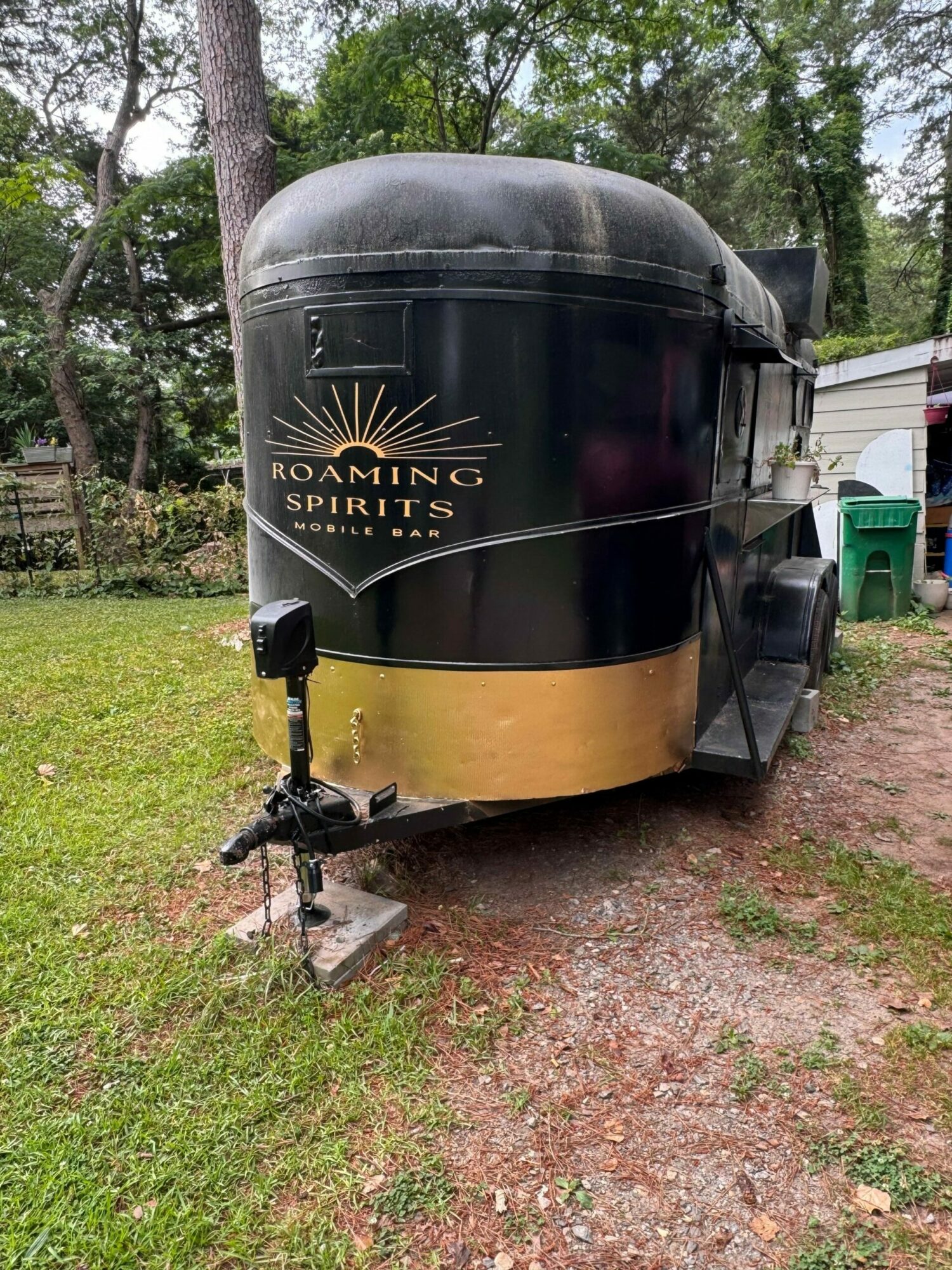 Black and gold trailer with a logo, parked on grass with trees in the background.