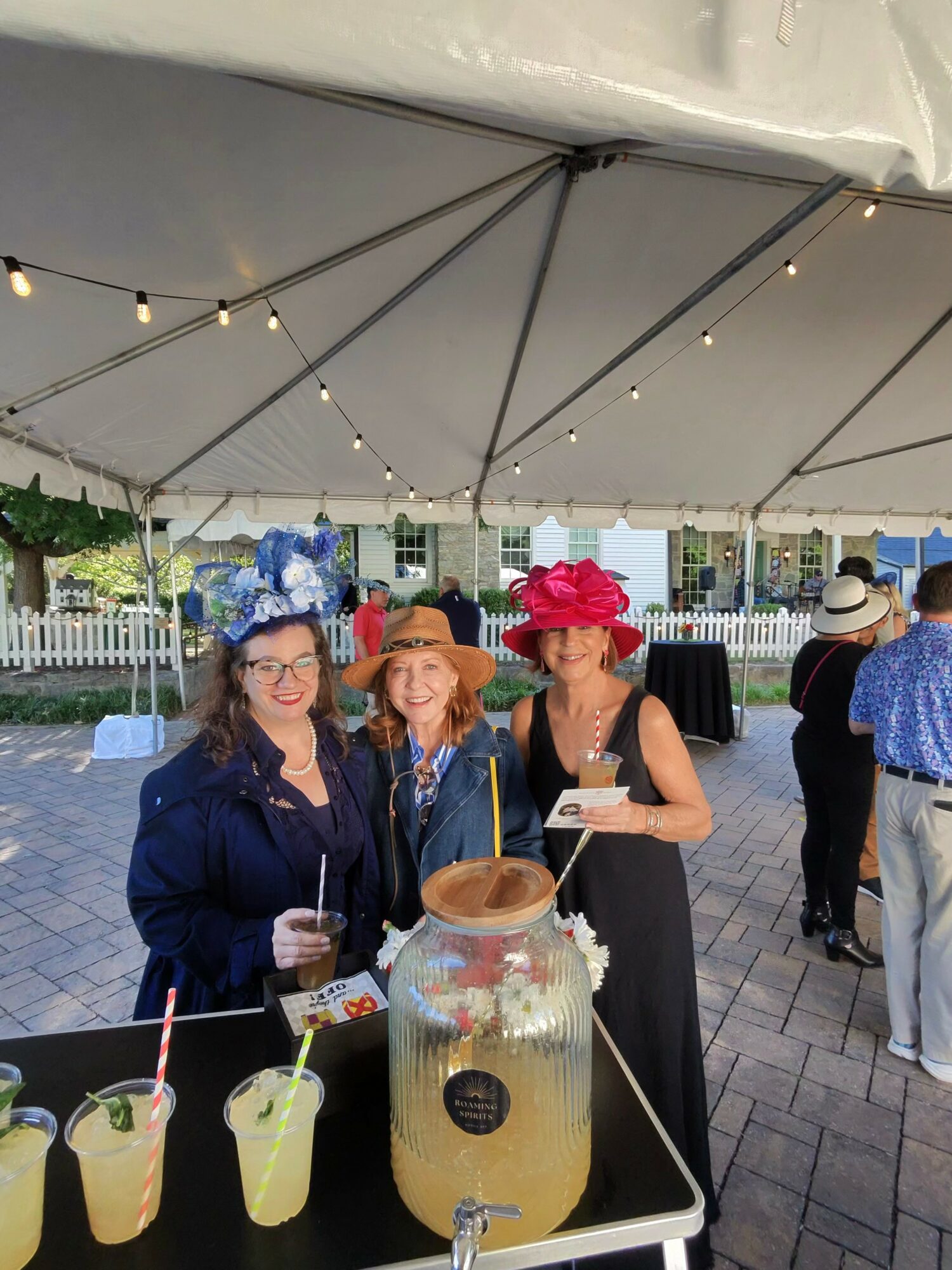 Three women in hats and dresses stand under a tent at an outdoor event, with drinks and a large jar on the table.