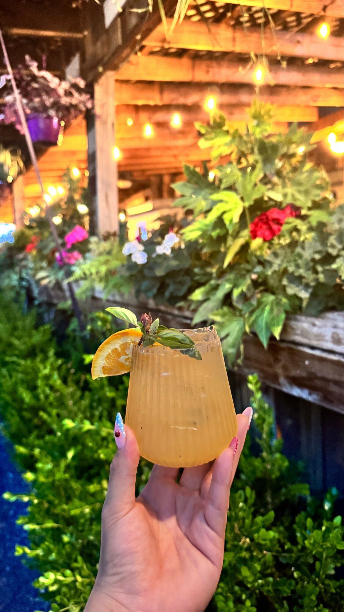 Person holding a pineapple-shaped drink with a lemon wedge in a garden setting with flowers and string lights.