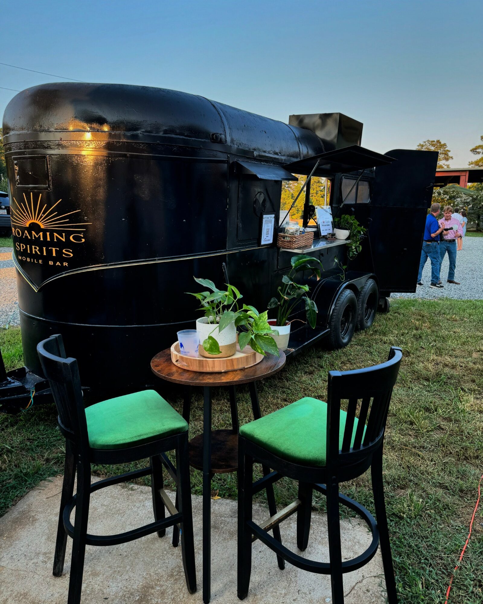 Black mobile bar with a logo, two green-cushioned chairs, a small round table with plants, outdoor setting with people in background.