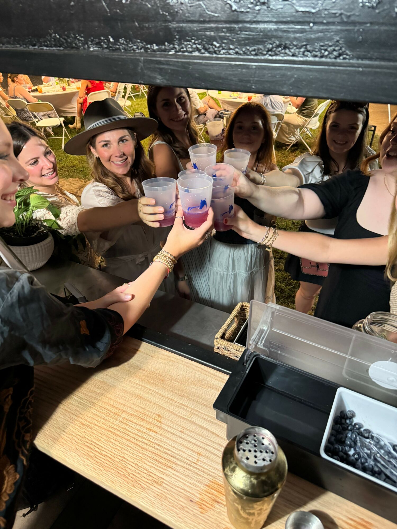 Group of women holding water bottles, some wearing hats, in a room with a wooden table and various objects.