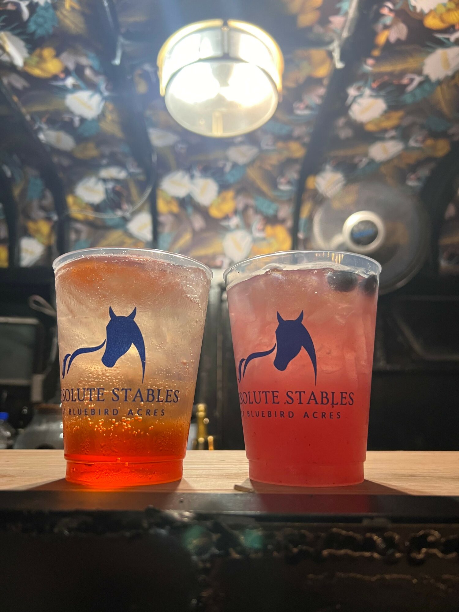 Two glasses of beer on a wooden surface, with a patterned background and a hanging light. The glasses have a blue logo and red liquid.