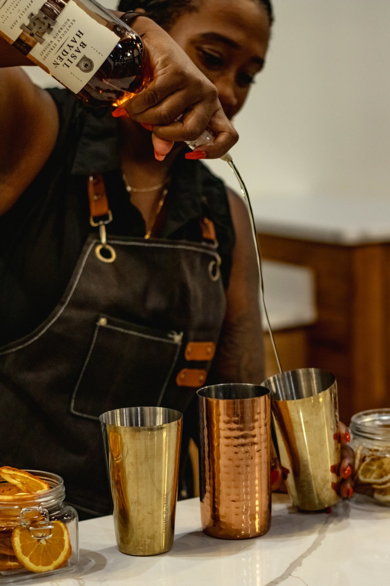 Person pouring a drink into a copper cup, with other cups and jars on a table, indoors.