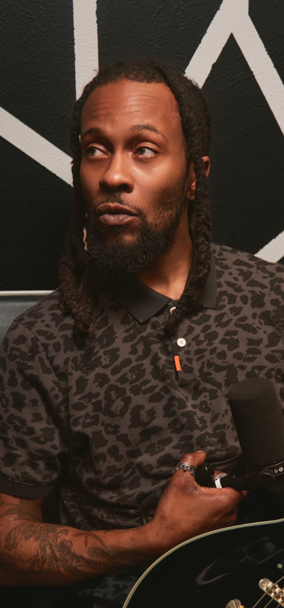 Man with long hair and beard wearing a patterned shirt, holding a microphone, standing against a dark background.