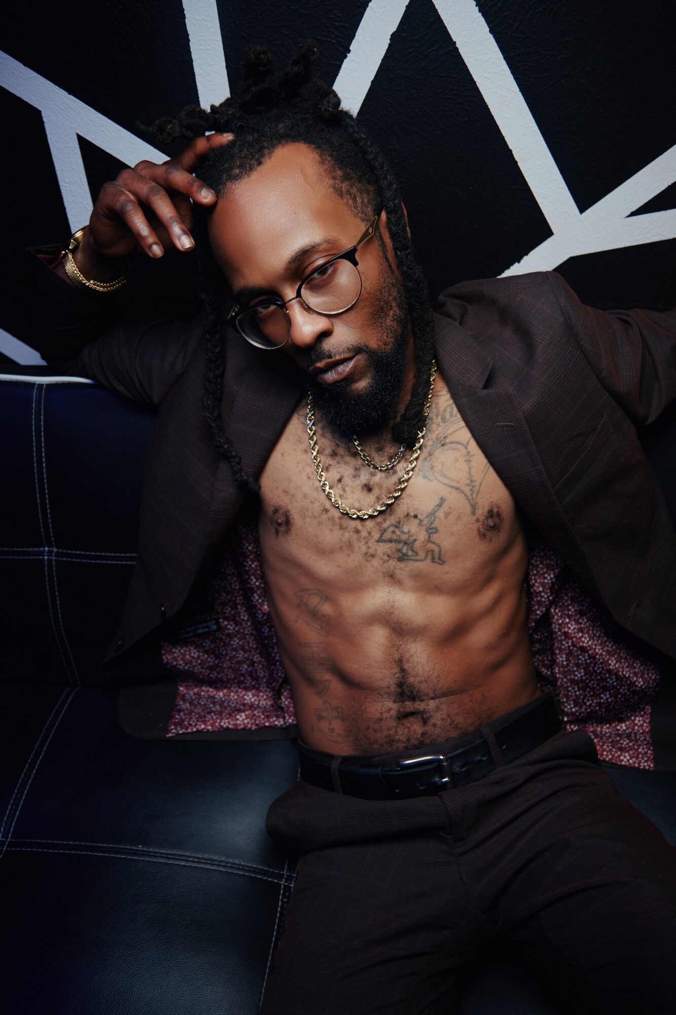 Man with glasses, dreadlocks, and tattoos, shirtless with open jacket, sitting against a geometric background.