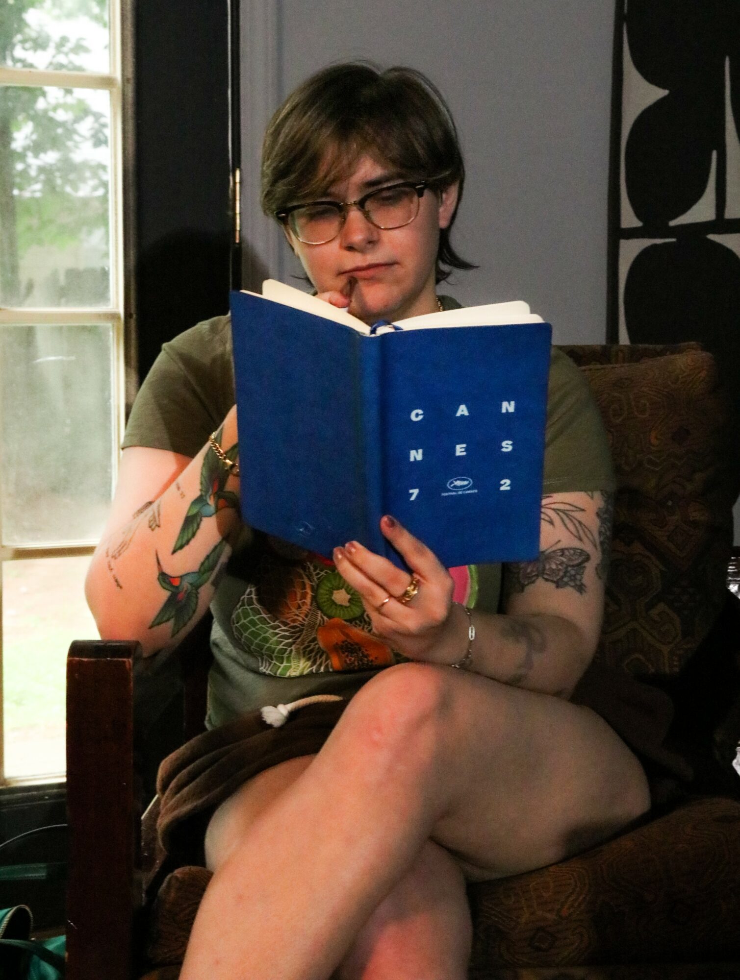 Person with short hair and glasses reading a blue book indoors, sitting on a dark-colored chair with tattoos visible on arms and legs.