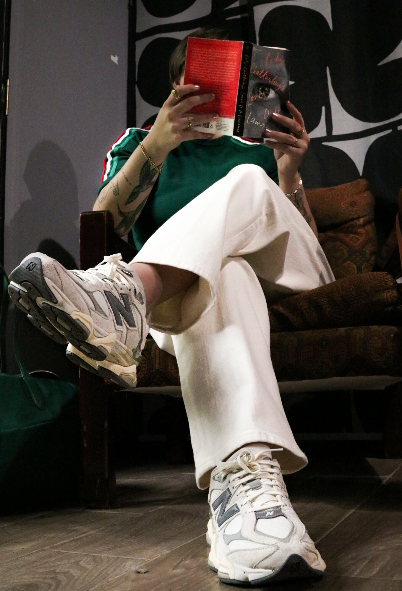Person sitting on a chair, holding a book in front of their face, wearing white sneakers and loose white pants.