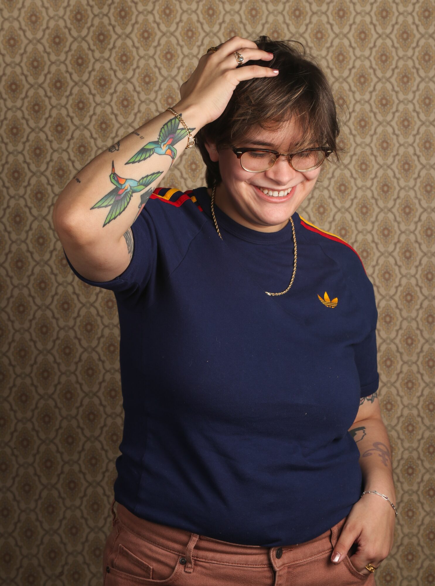 Person with glasses smiling, hand on head, wearing a navy shirt with a small yellow logo, standing against patterned background.