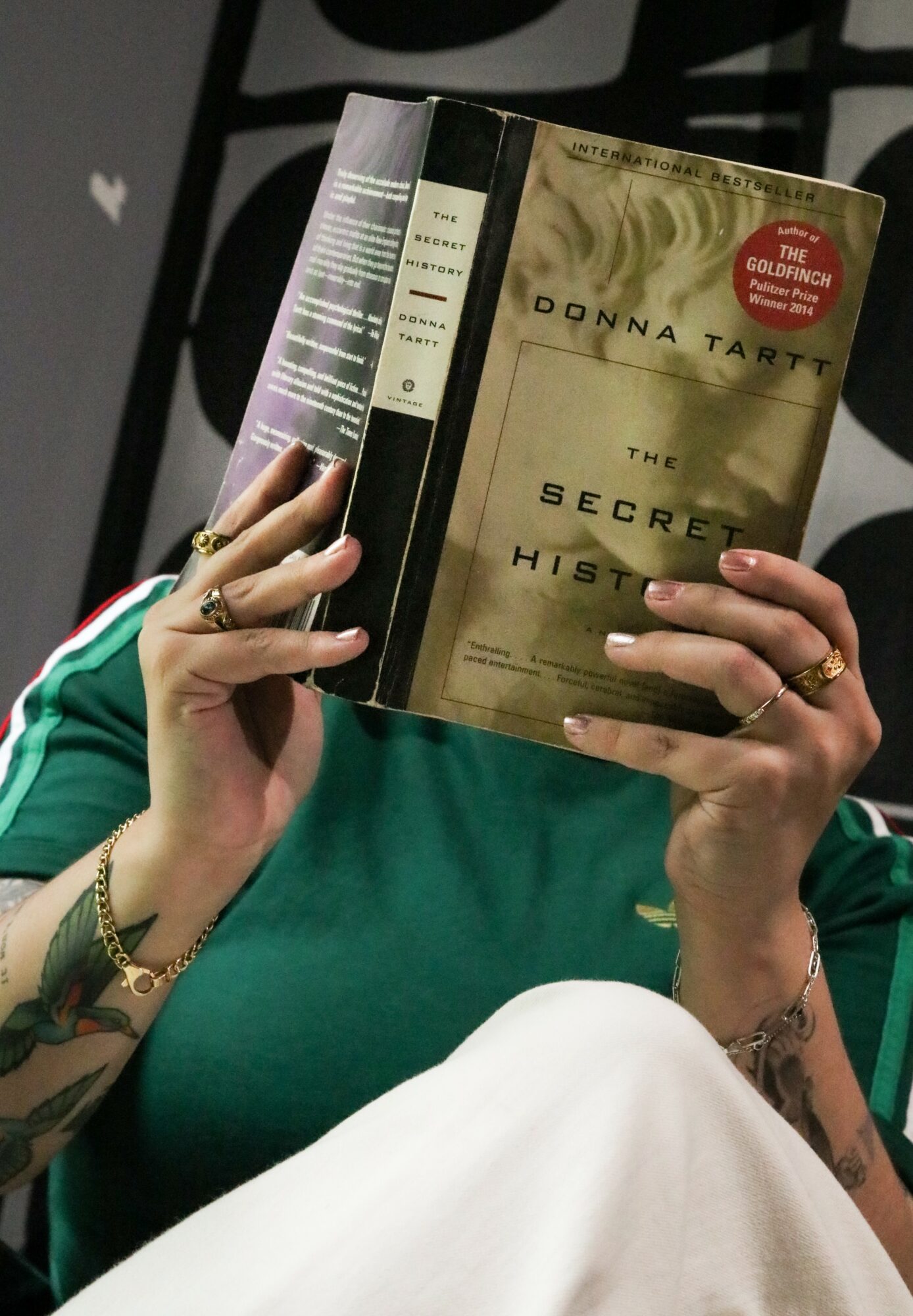 Person holding a book titled 'The Secret History' by Donna Tartt, covering their face, wearing a green top and rings.