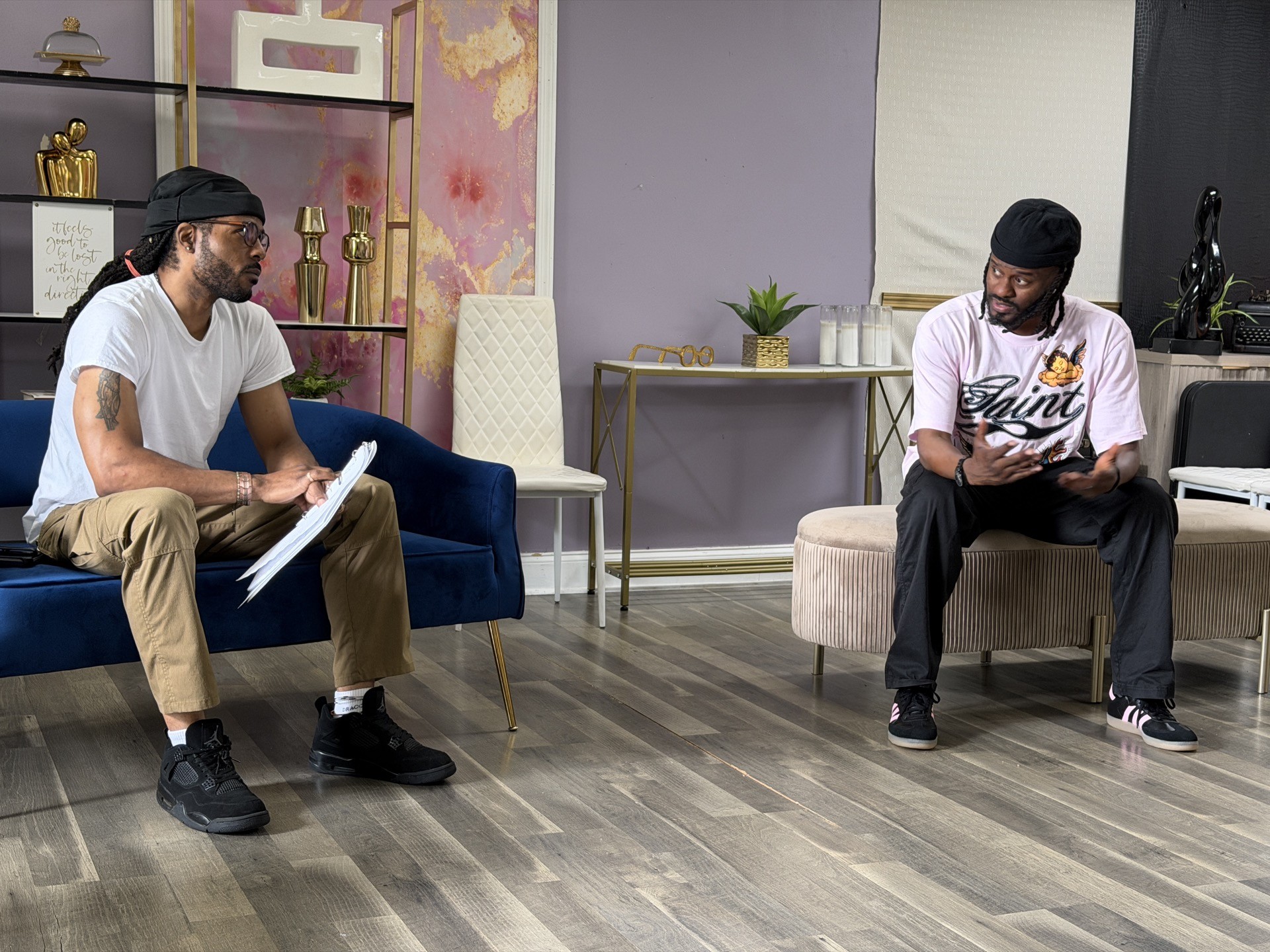 Two men sit on chairs in a room with colorful wall panels, one holding a notebook and the other a phone.