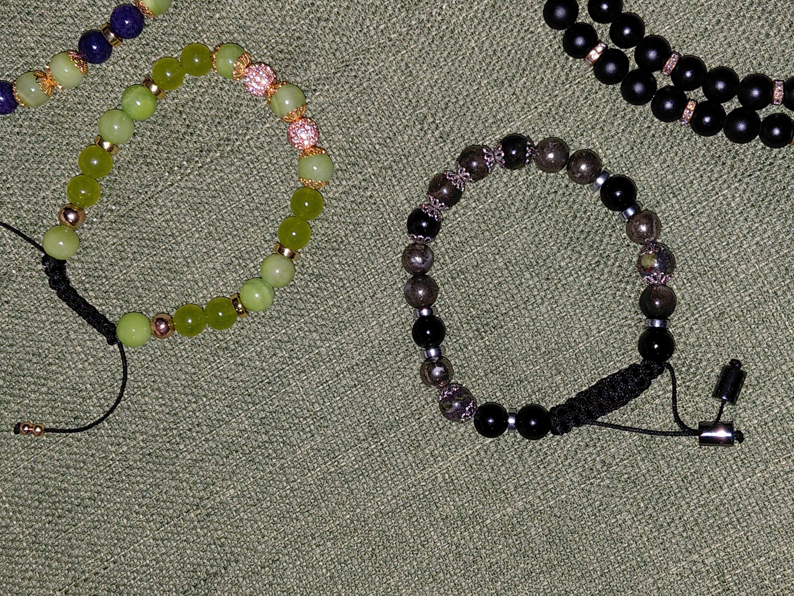 Two beaded bracelets on a textured surface, one with green, pink, and white beads, the other with purple and black beads.