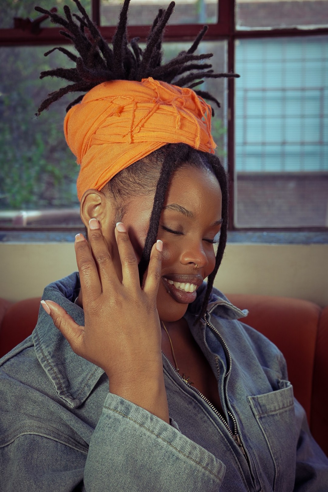 Woman with dreadlocks wearing an orange headwrap, smiling with eyes closed, in a casual setting.