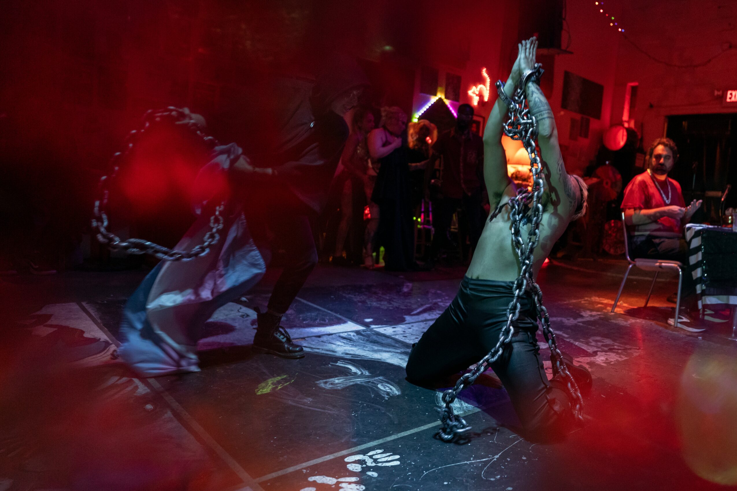 Person performing a dance with chains in a dimly lit room with a red hue, audience in background, some seated.