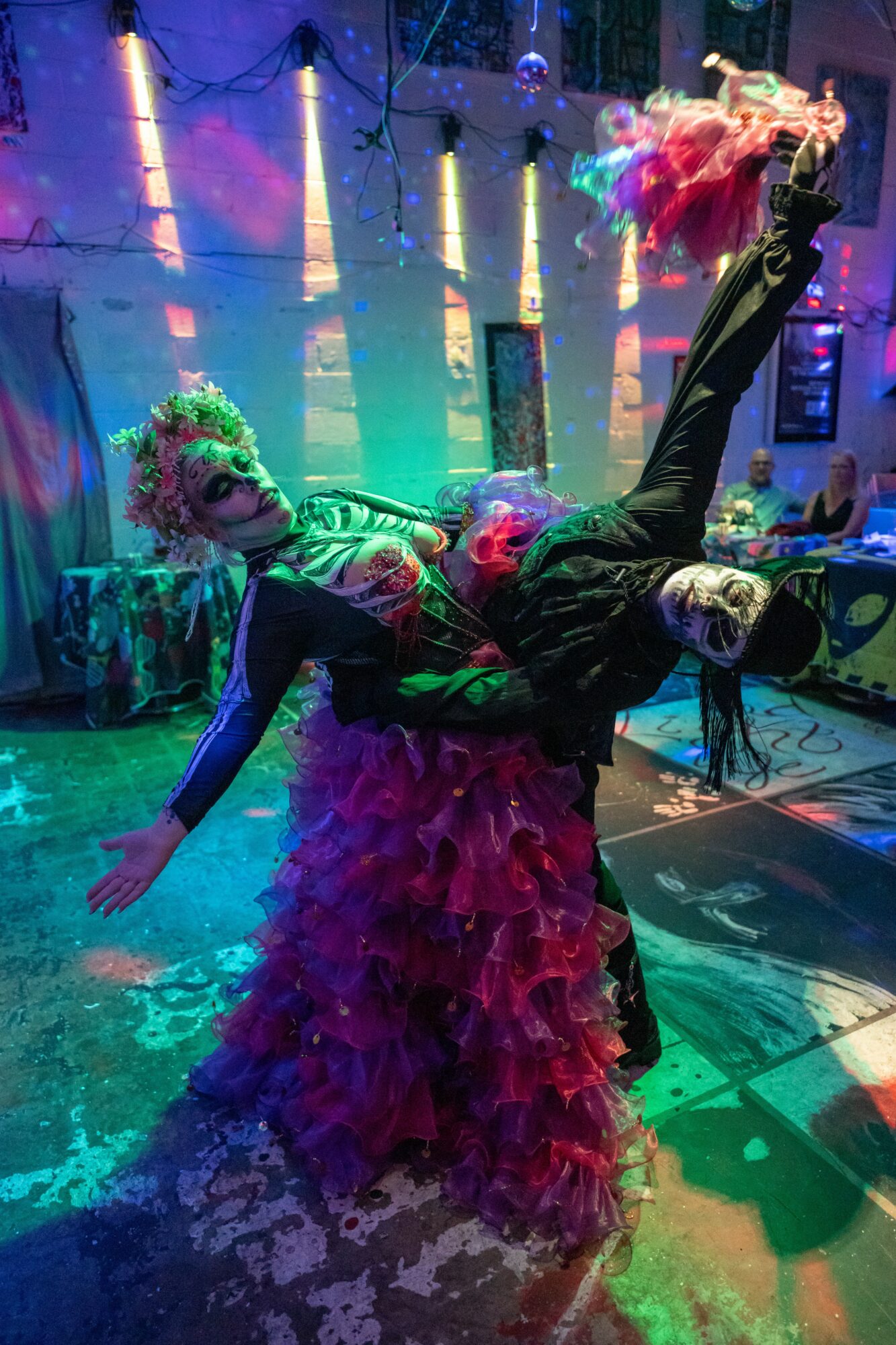 Two performers dance on stage with colorful lighting, one in a ruffled dress and the other in dark clothing, holding a fabric.