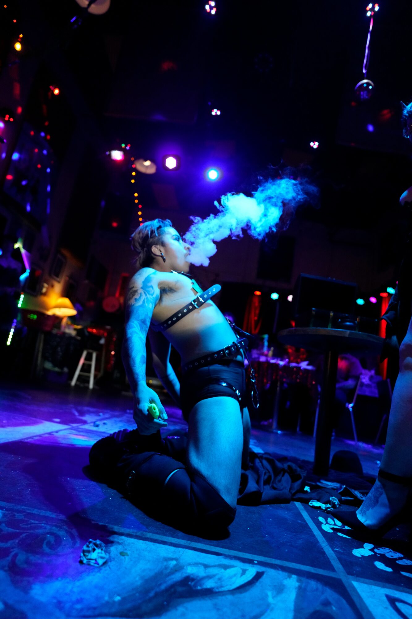 Person kneeling on stage, exhaling vapor, with colorful lights and a dark background, performing in a nightclub or concert setting.