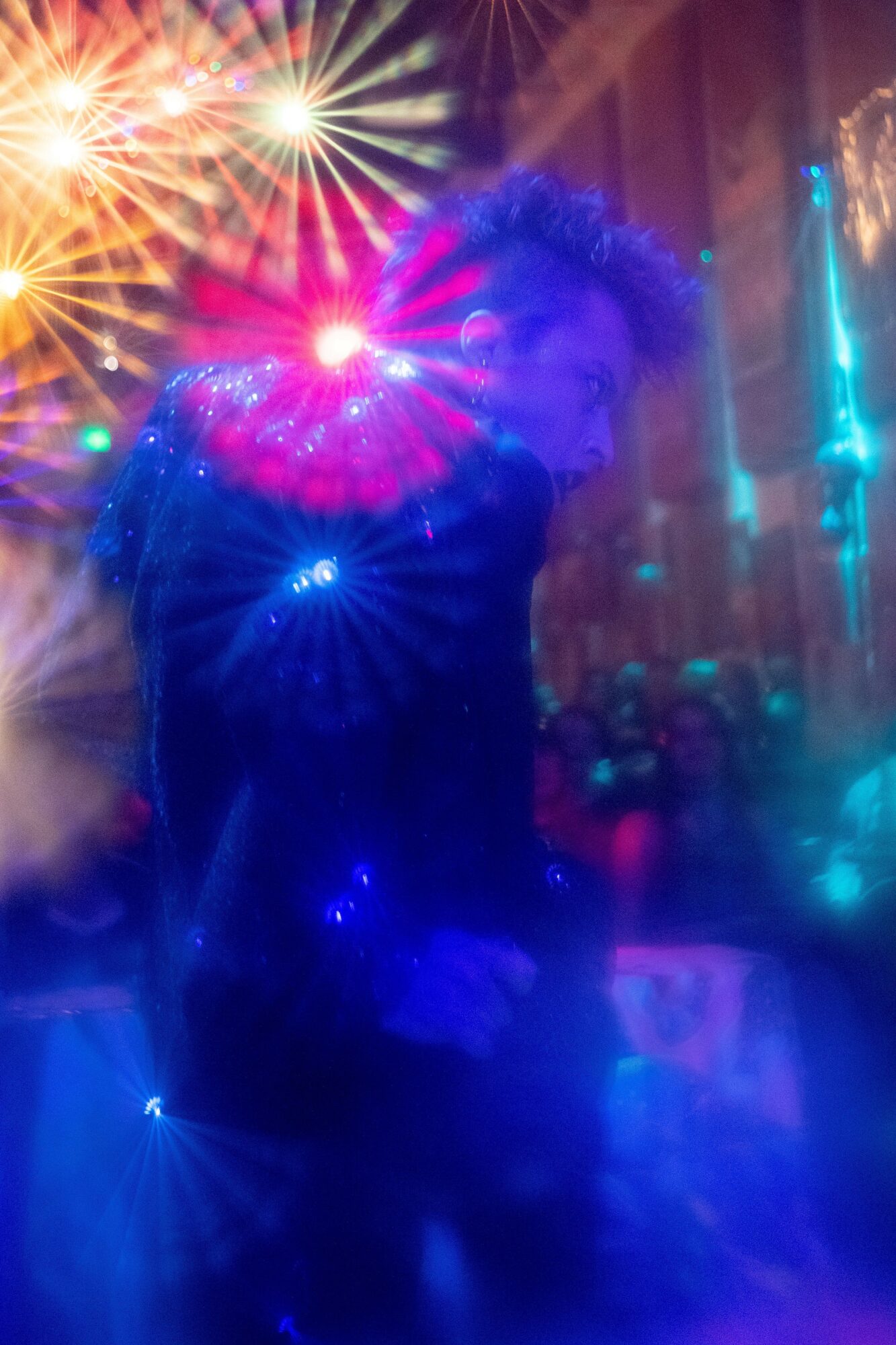 Person standing in a colorful, illuminated environment with bright lights and reflections, facing slightly to the side.