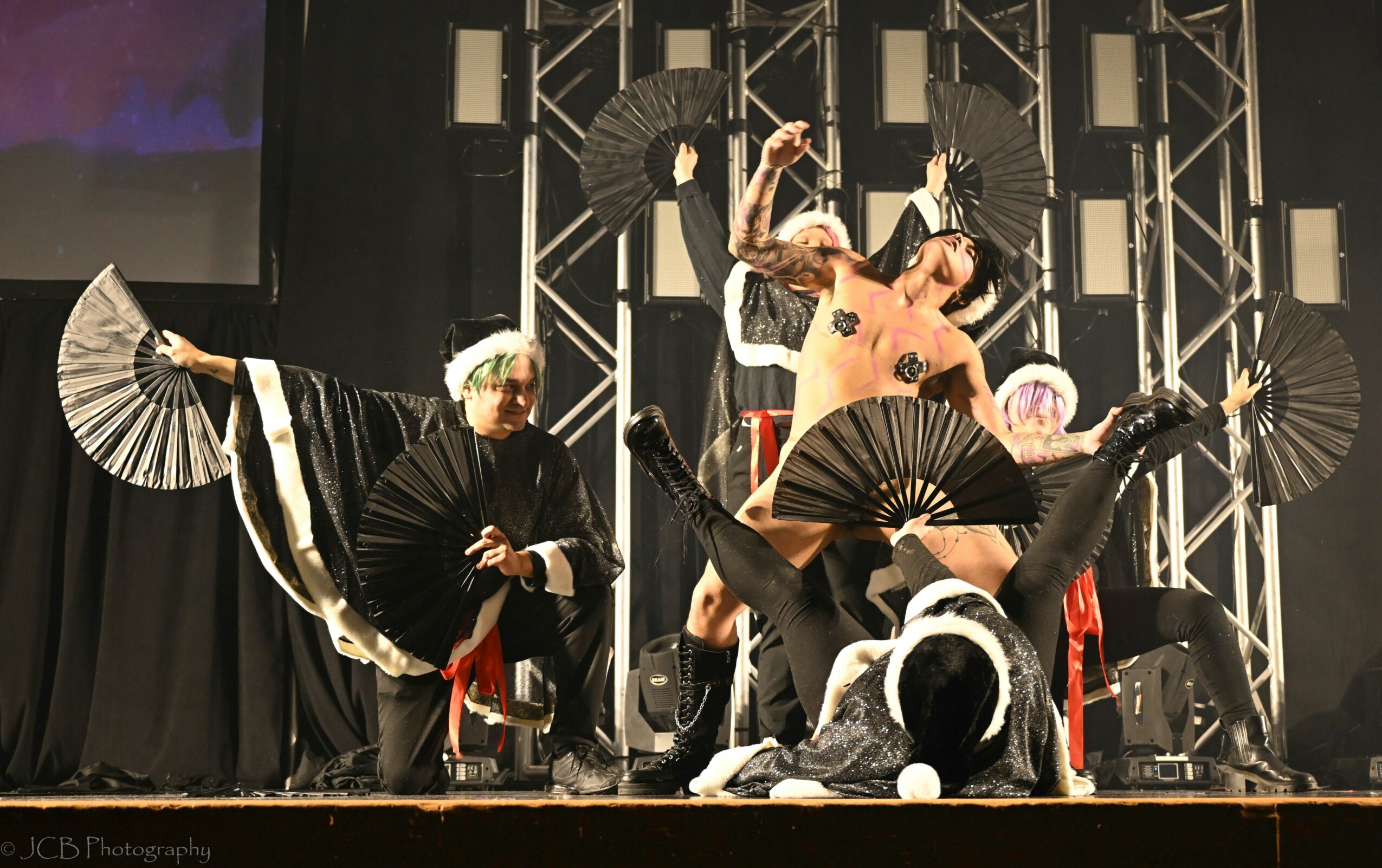 Group of performers on stage with fans, some in costumes, performing a dance or act, with stage lighting and scaffolding.